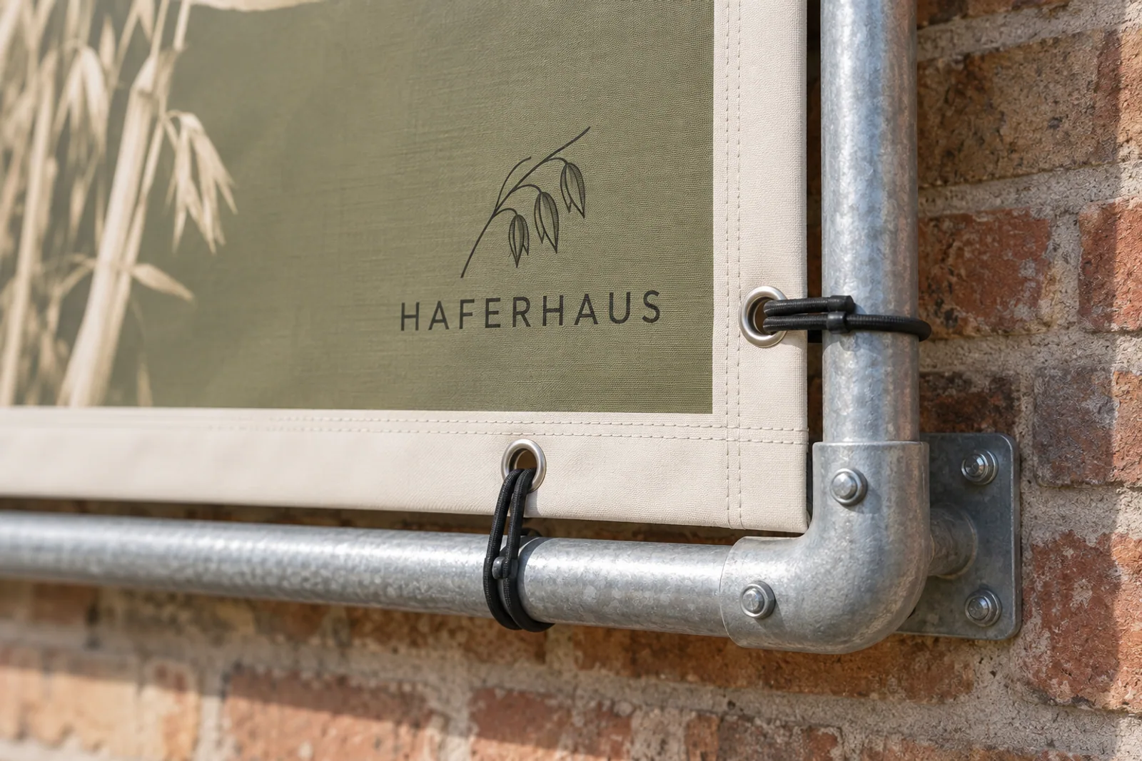 Verschraubte Rohrrahmenecke mit Spannschlaufen, Metallösen und verstärktem Saum eines olivgrünen HAFERHAUS-Banners vor einer Backsteinwand.