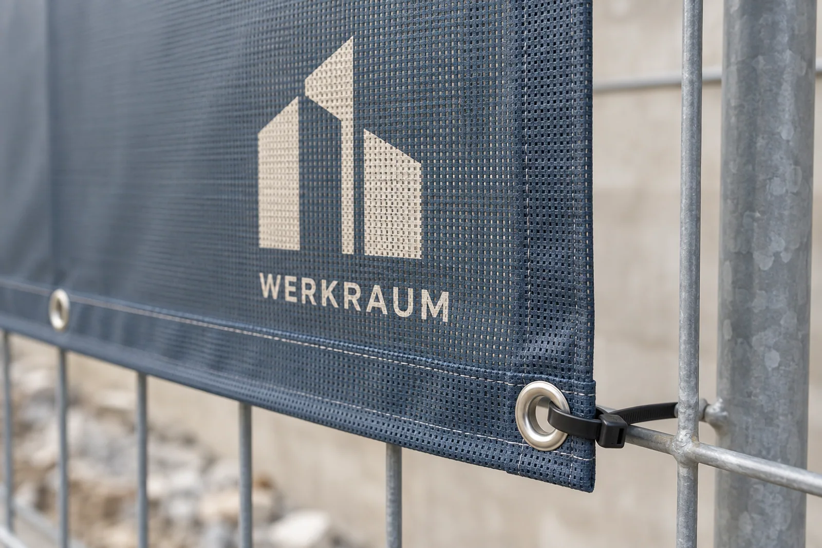 Nahaufnahme eines blau bedruckten Mesh-Banners „WERKRAUM“ mit verstärktem Saum, Metallöse und schwarzem Kabelbinder am verzinkten Bauzaun.