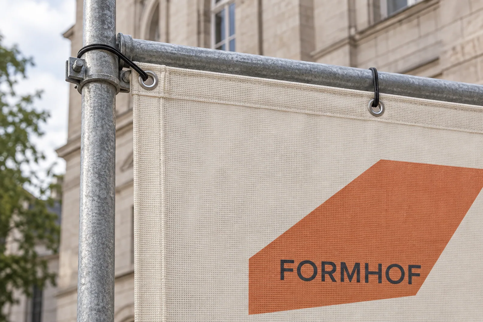 Verstärkte Ecke eines beigefarbenen FORMHOF-Meshbanners mit Metallösen und schwarzen Spannschlaufen an verzinkten Gerüstrohren.