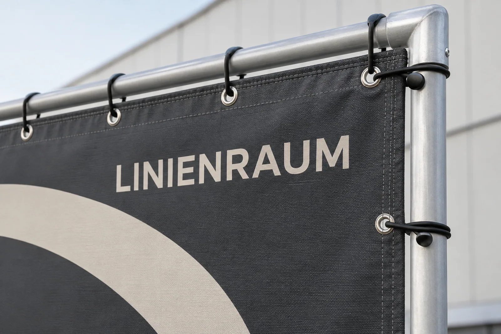 Detail eines anthrazitfarbenen Großformatbanners „LINIENRAUM“ mit verstärkter Kante, Metallösen und schwarzen Spannschlaufen an einem Aluminiumrahmen.