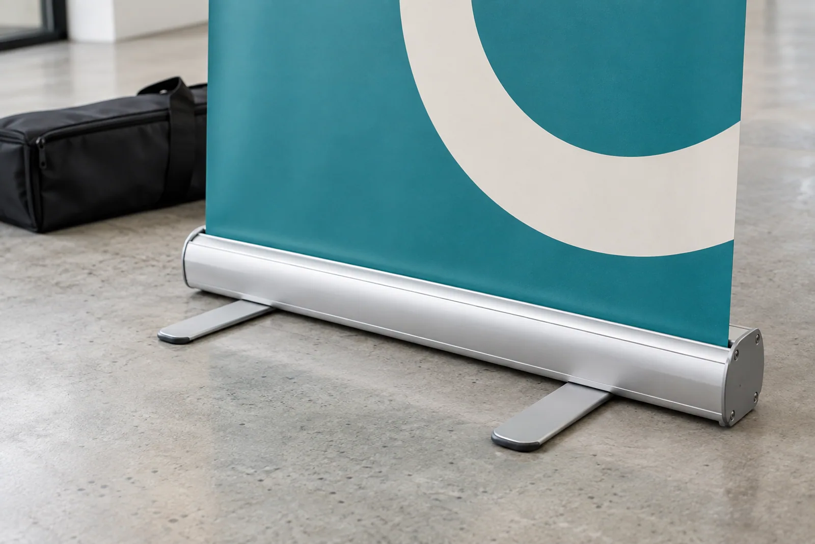 Silberne Roll-up-Kassette mit zwei Standfüßen und türkisfarbener Grafikbahn auf einem Betonboden.