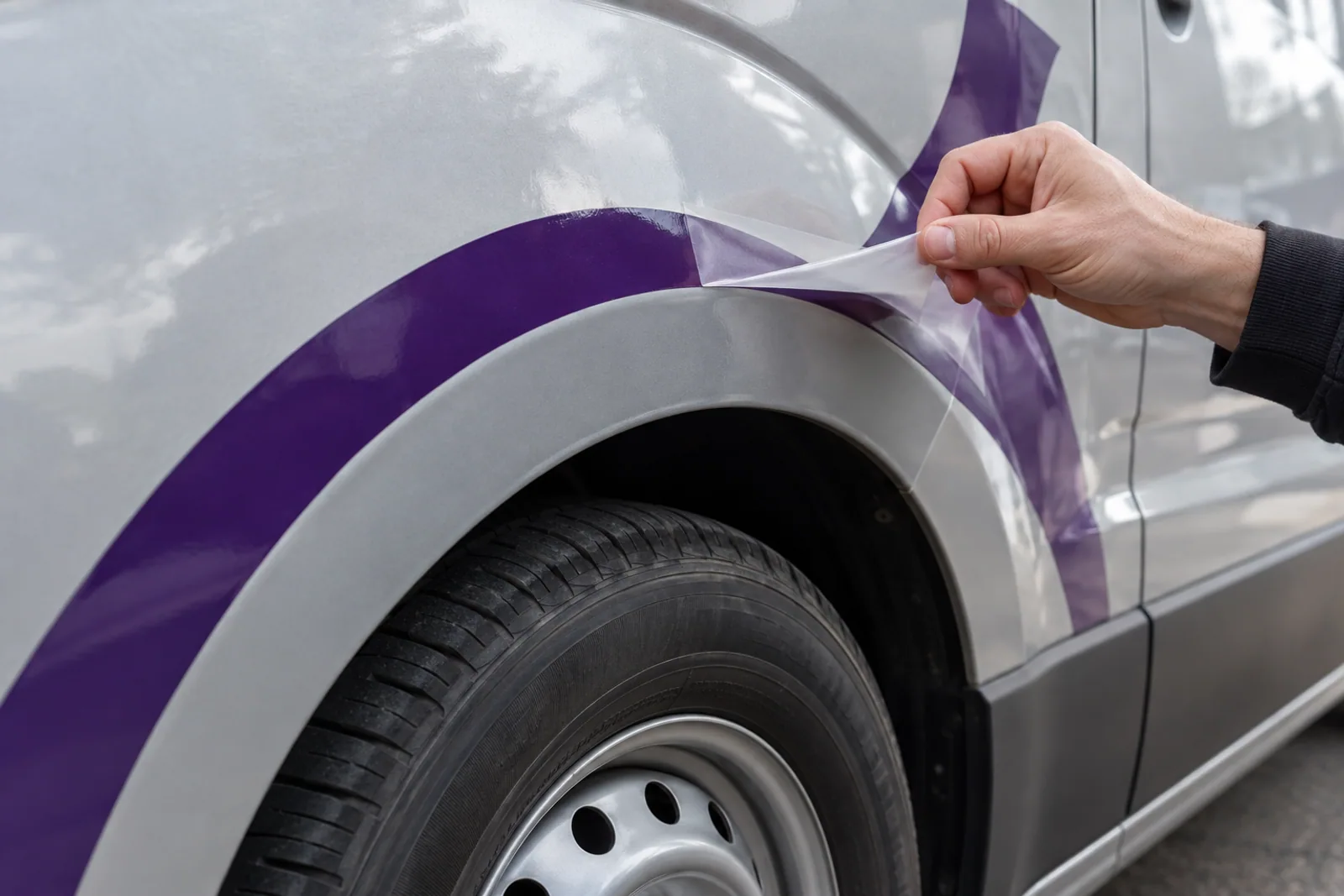 Eine Hand zieht Transferfolie von einer violetten Grafik entlang des Radlaufs eines silbernen Transporters ab.