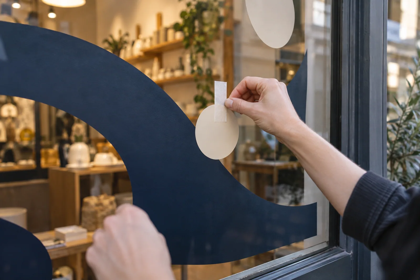Eine Hand positioniert ein helles Kreismotiv mit Montageband auf einer blau gestalteten Schaufensterscheibe.