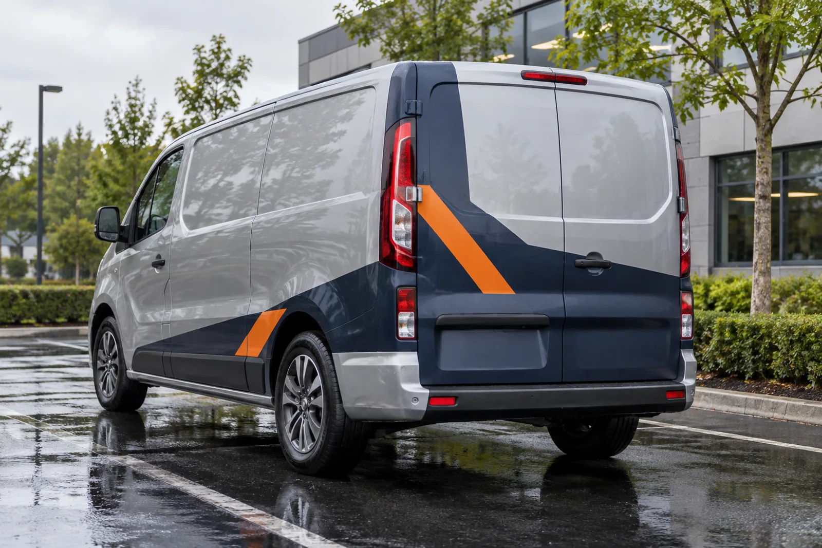 Silberner Transporter mit dunkelblauer und orangefarbener Teilfolierung in der Heckansicht.